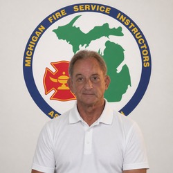 Portrait of a man in a white polo shirt standing before the Michigan Fire Service Instructors emblem featuring a green Michigan map and a red fire cross.