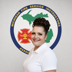 Portrait of a smiling woman in a white polo shirt standing in front of the Michigan Fire Service Instructors Association logo plate on the wall behind her.