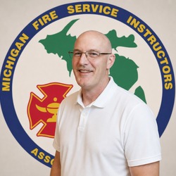 Portrait of a smiling, bald man with glasses in a white polo shirt, standing in front of the Michigan Fire Service Instructors Association logo.