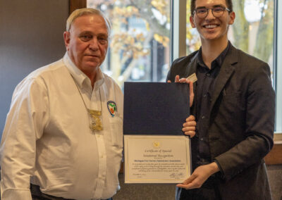 Two men smile for a photo; older man in a white shirt with badges beside a younger man in a dark suit holding a certificate folder.
