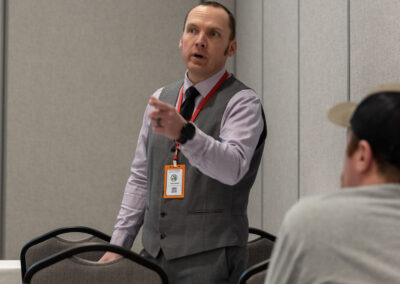 Man in a gray vest and red lanyard speaking and gesturing in a conference room with chairs and tables nearby.