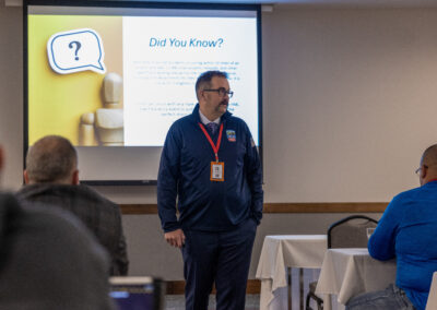 Male presenter in blue uniform with a red lanyard speaking in a conference room, slide reads 'Did You Know?' behind him.