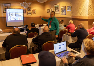 Man in teal shirt presents a slide show to an audience in a conference room with a projector screen in front of them.
