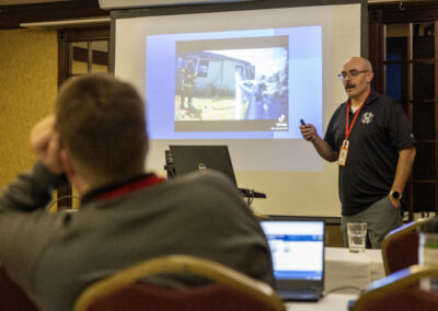 Male presenter with mustache in dark polo and red lanyard presenting a slide to an audience in a conference room.