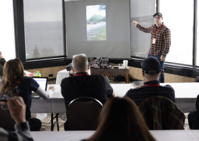 Presenter in a plaid shirt and cap points to a projection screen at the front of a meeting room, attendees seated and watching.