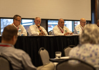 Panel of five men in white shirts seated behind a long table, one speaking into a microphone; conference setting indoors with water bottles on the table