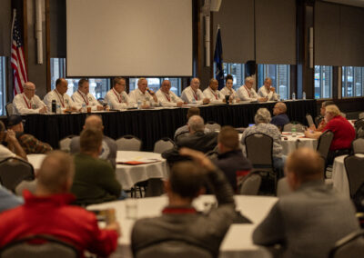 Panel of speakers in white shirts seated behind a long table at a conference, audience in foreground; American flag visible on left.