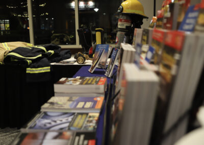 Display table featuring firefighter gear (yellow helmet, turnout coat, breathing apparatus) with brochures and magazines in a dimly lit setting, shelves blurred in the foreground.