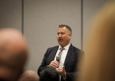 Middle-aged man in a dark suit and tie speaking into a handheld microphone at a conference, with blurred attendees in the foreground.
