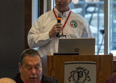 Speaker in a white shirt with a microphone at a podium, presenting at a hotel conference center.