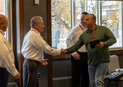 Four men in business casual shake hands in a room near large windows with autumn leaves outside.