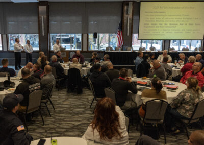 Conference room full of attendees at round tables, presenting slides on screen with an American flag nearby and two speakers at the front.