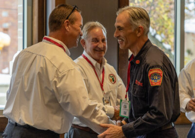 Three men in uniform shirts shake hands in a doorway at an indoor event; badges on lanyards visible.