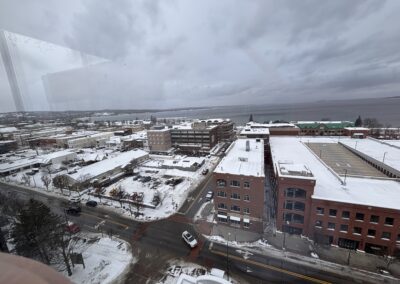 Snowy urban riverfront viewed from above, with buildings, streets, and a cloudy sky beyond the water's edge.