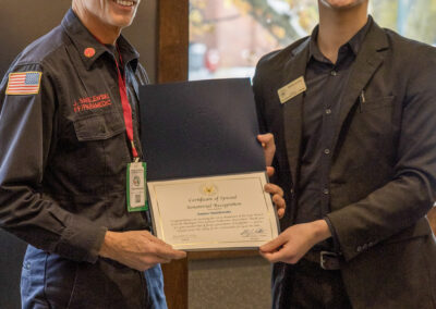 Two men standing indoors, one in a uniform presenting a certificate to a smiling younger man in a dark blazer and glasses.