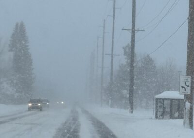 Snowy street with falling snow, utility poles and power lines lining the road; cars with headlights approaching in the distance.