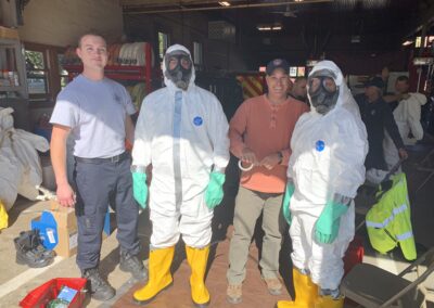 Group of people in hazmat suits and protective gear posing in a fire station, with hoses and equipment in the background.