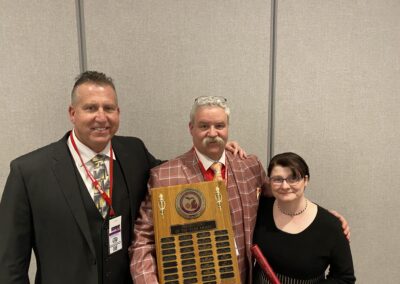 Three people posing with a tall multi-plate wooden award plaque at a conference or event.