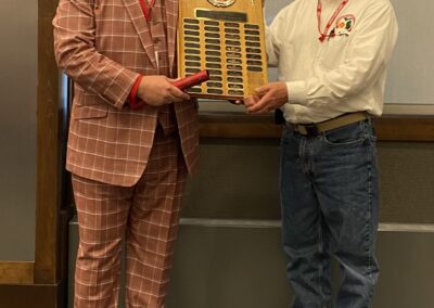 Two men pose in a hallway while holding a large wooden award plaque with many nameplates.