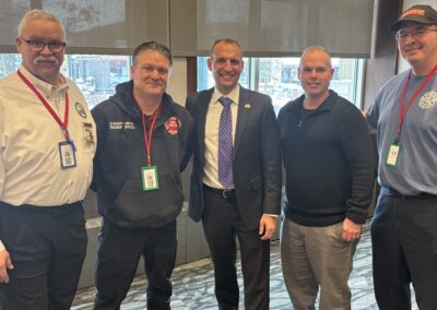 Five men in a line posing for a photo indoors; two wear badges, one in a suit, others in casual work attire, smiling at camera.
